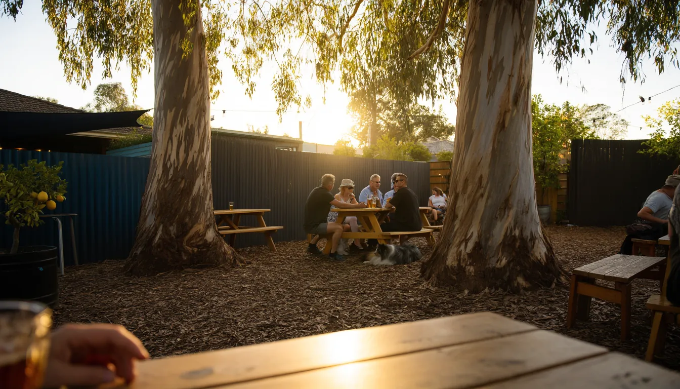 A suburban pub beer garden late on a summer afternoon, a group around a picnic table under two big gums with a dog lying in the bark chips beside them.