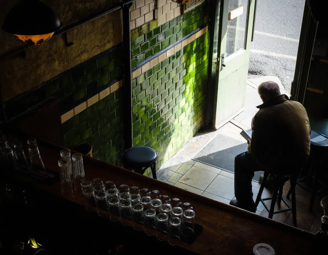 An old tiled corner pub front bar at eleven in the morning, shot from behind the counter past a row of clean upturned glasses, with one customer down the far end.